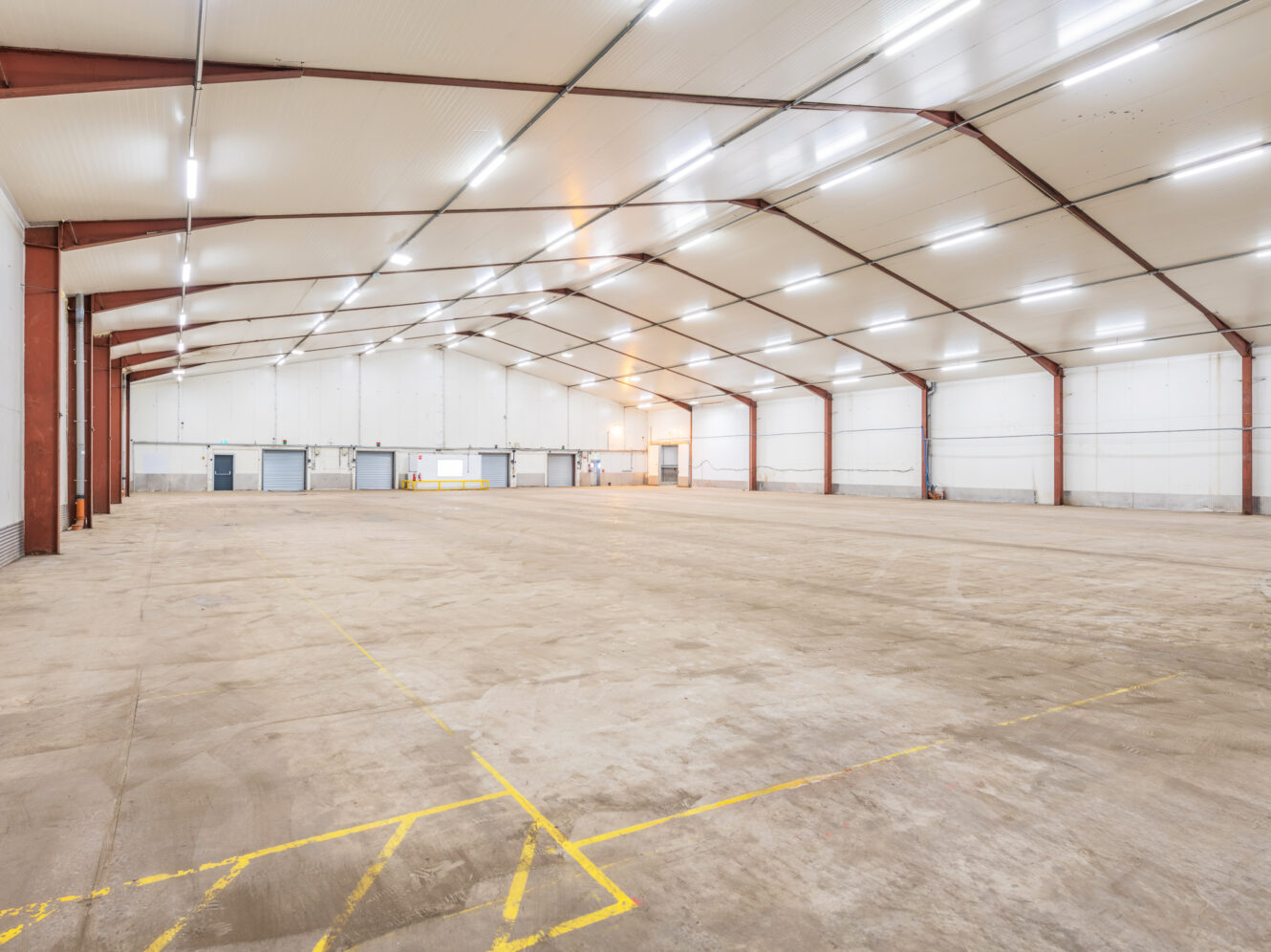 Large, empty warehouse with high ceilings, exposed beams, white walls, concrete floor, and bright overhead lighting. Yellow lines are marked on the floor near the entrance.