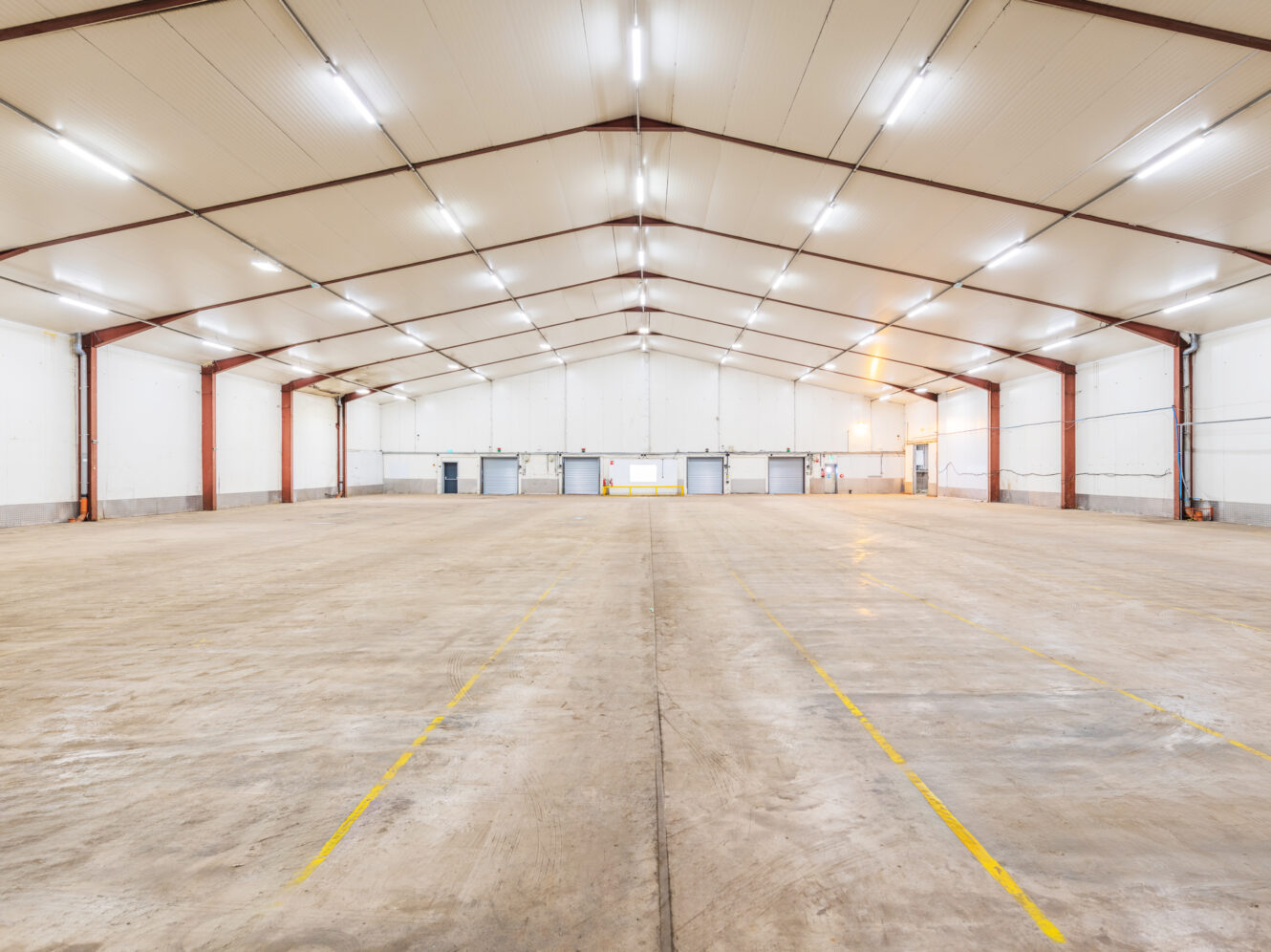 Wide-angle view of an empty warehouse with high ceilings, overhead lighting, concrete floor, and multiple large loading doors at the far end.