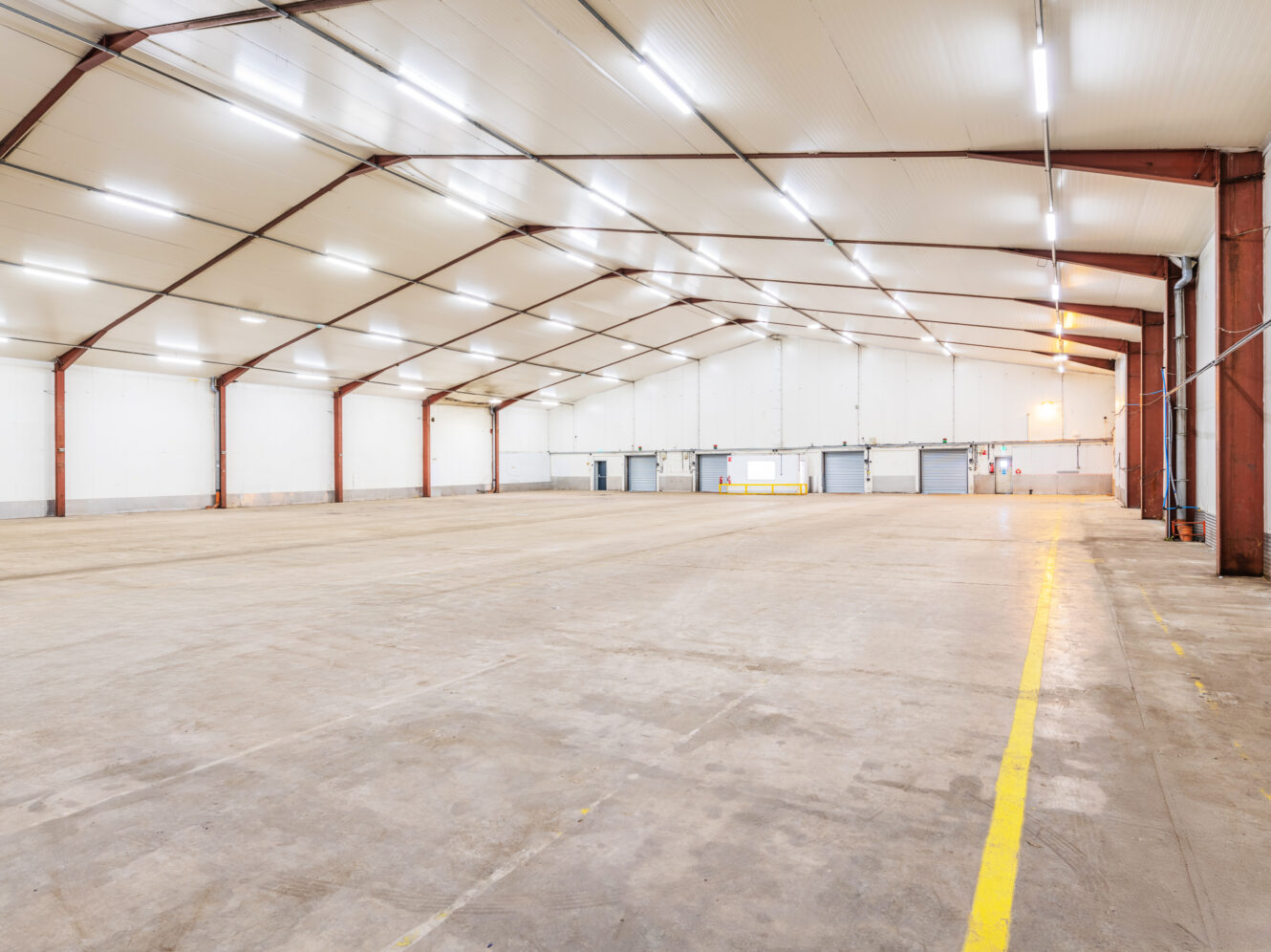 Spacious, empty warehouse with a high ceiling, exposed beams, concrete floor, white walls, and overhead fluorescent lighting. Yellow lines mark areas on the floor.