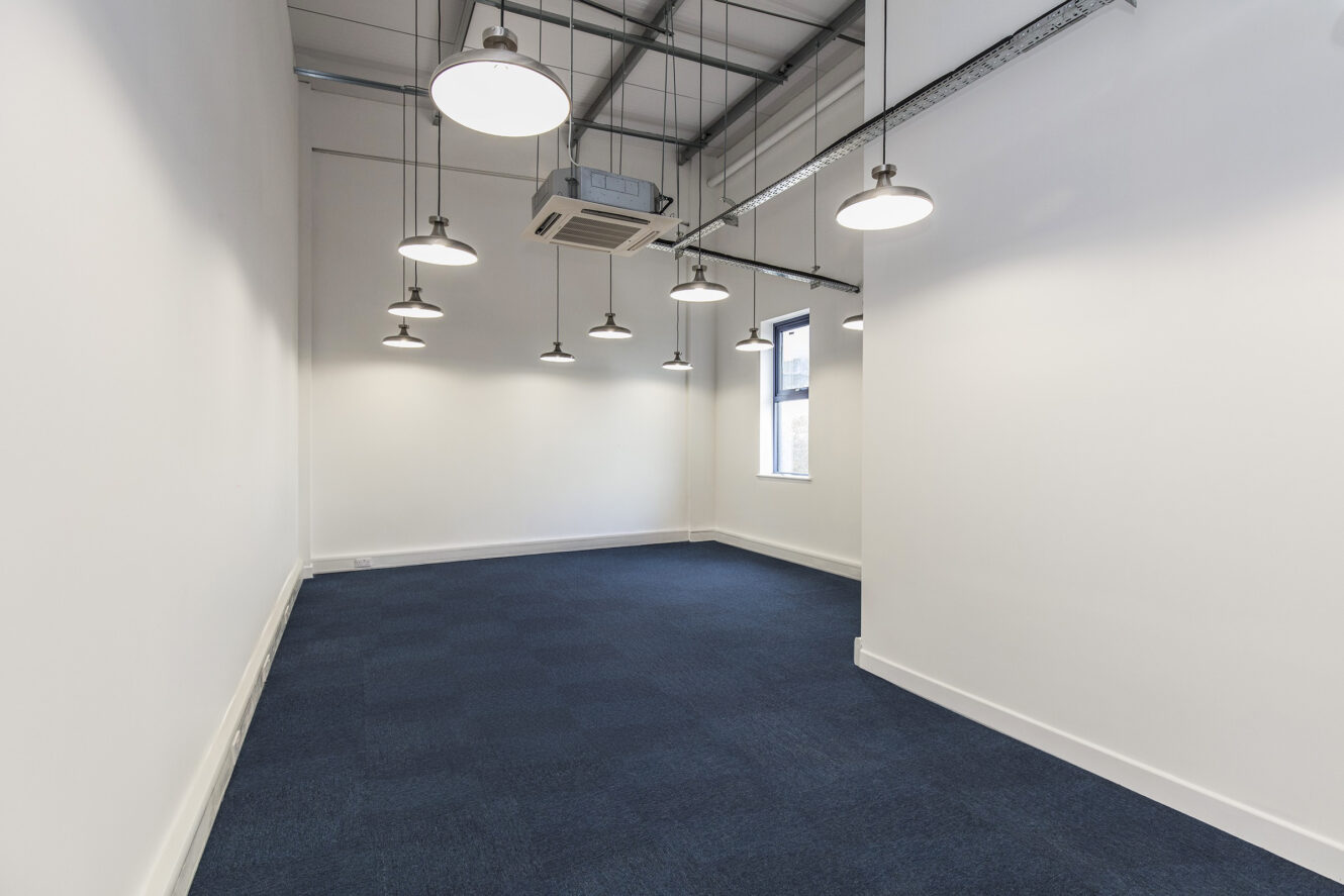 Empty office space with blue carpet, white walls, several ceiling lights, exposed ductwork, and a window on the right wall.