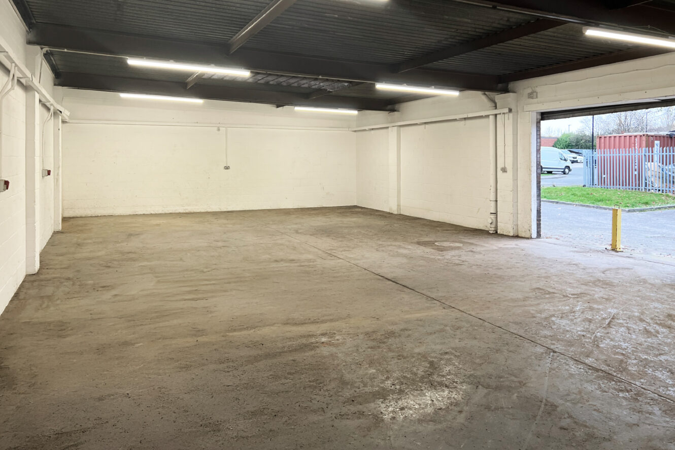 Empty industrial warehouse with concrete floors, white brick walls, exposed ceiling beams, and fluorescent lighting, with an open garage door showing a view outside.