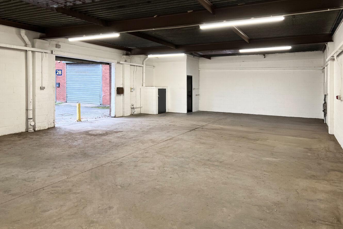 Empty industrial warehouse unit with concrete floor, white-painted brick walls, exposed beams, and an open roller shutter door leading outside.