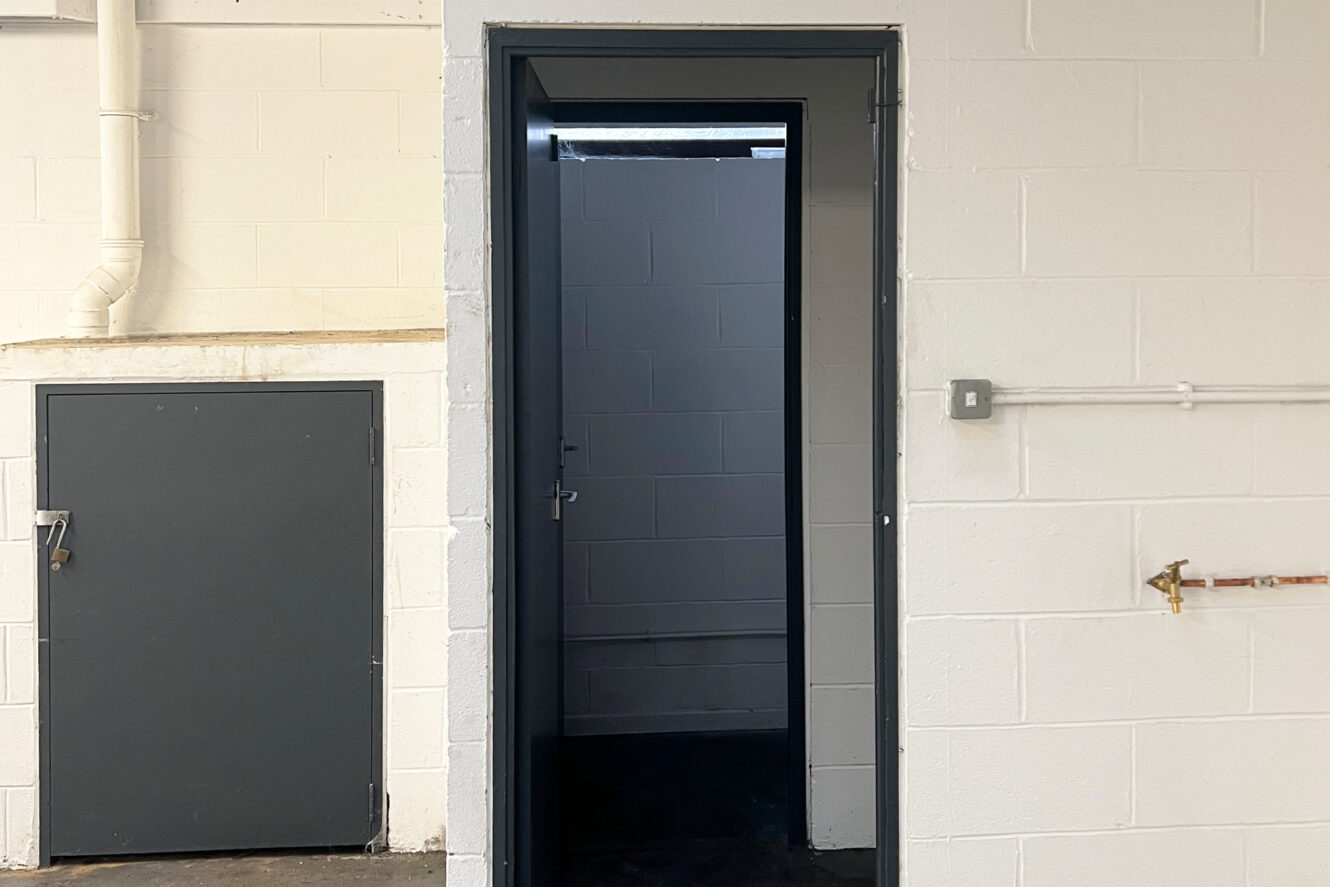 A gray metal door is open in a white brick wall, revealing a dimly lit corridor with another closed door inside. Pipes and an electrical switch are visible on the surrounding walls.