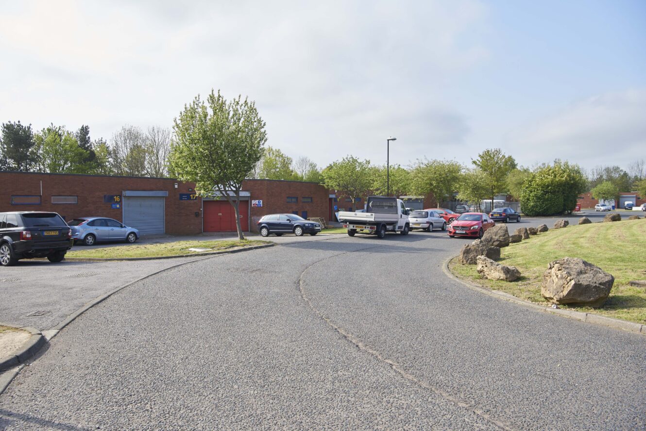 A curved road with several parked and moving cars, bordered by grass, rocks, and brick buildings under a partly cloudy sky.