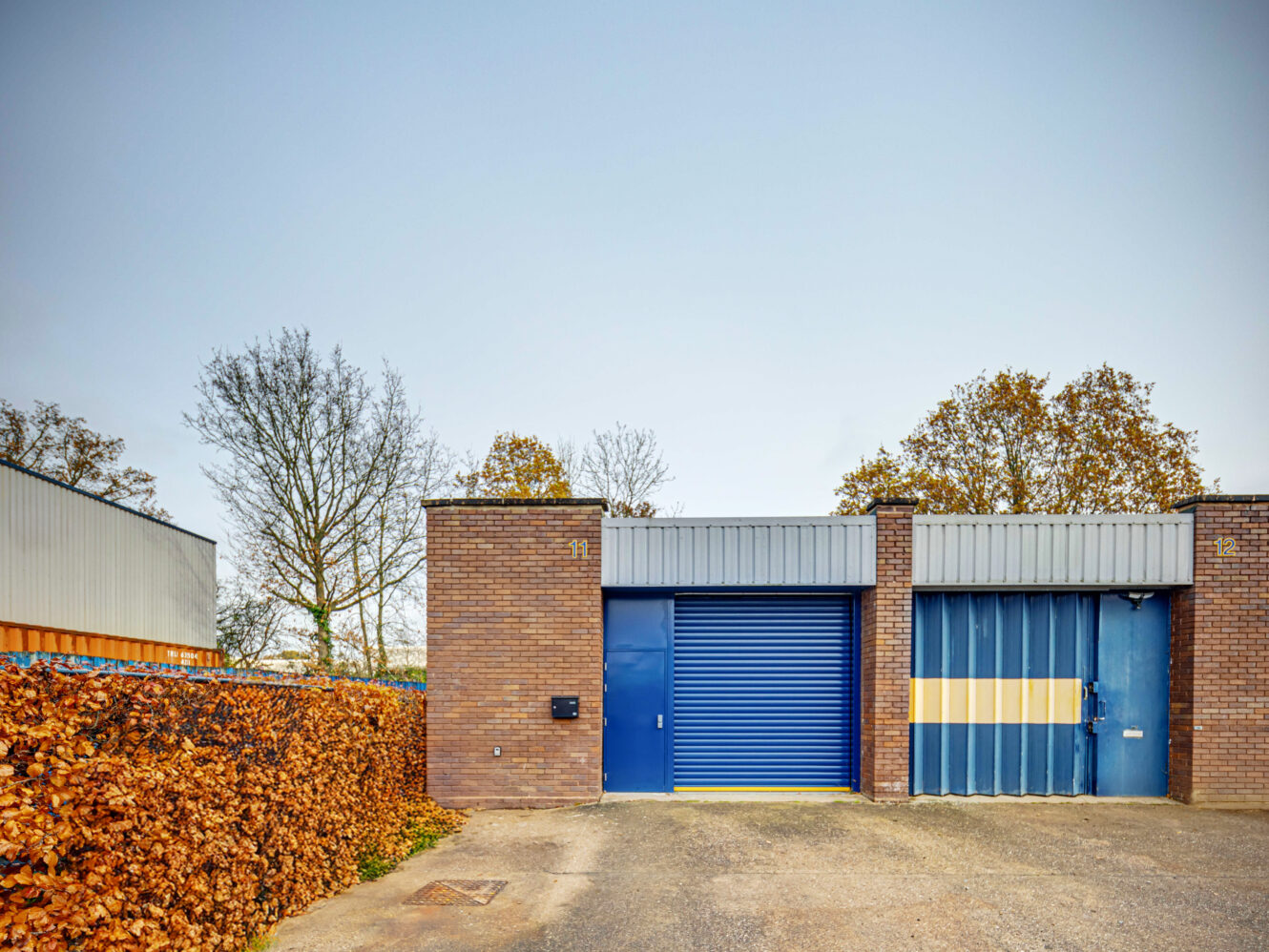 A brick industrial building with two blue garage doors and a hedge on the left side, photographed under a clear sky.