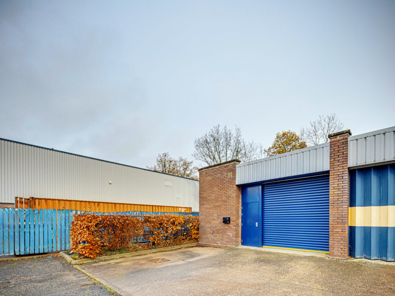 Industrial building with a blue roller shutter door, brick wall, and small hedge in front; adjacent to a corrugated metal fence and another building. Overcast sky in the background.