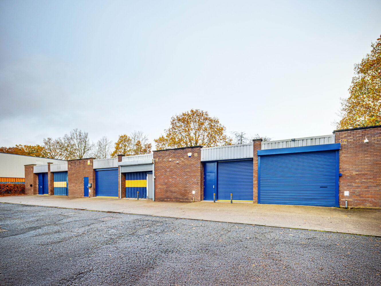 Row of brick industrial storage units with closed blue roller shutter doors, situated on an empty paved lot, with trees and cloudy sky in the background.