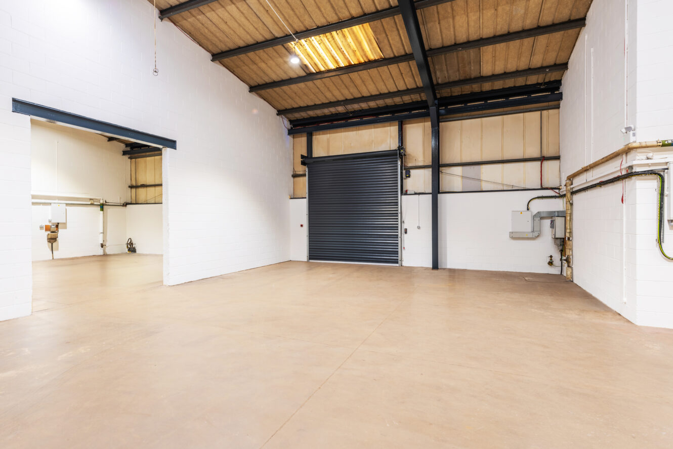 Spacious empty warehouse interior with white walls, high ceilings, concrete floor, overhead lights, and a large closed roller shutter door.