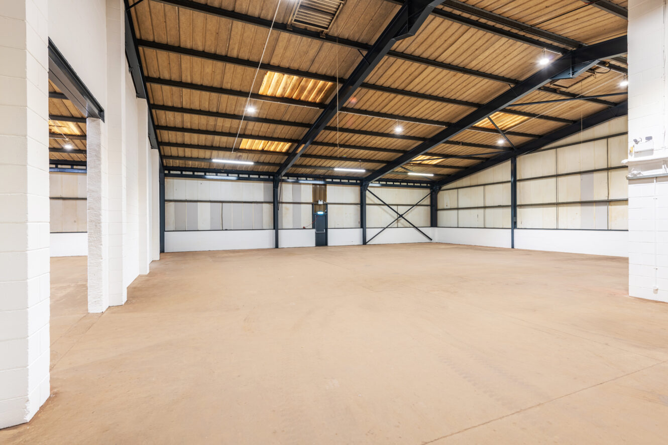Empty industrial warehouse interior with concrete floor, white painted walls, exposed metal beams, and a closed door at the back. Fluorescent lights are mounted on the ceiling.