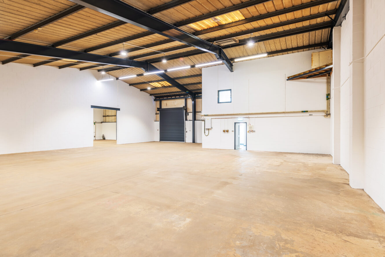 Spacious, empty warehouse with high ceilings, exposed beams, white walls, concrete floor, and large industrial door.