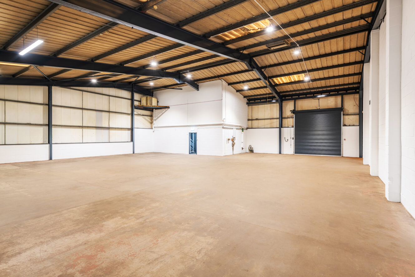 Empty warehouse interior with concrete floor, white walls, exposed metal ceiling beams, shelving, a blue door, and a large roller shutter door. Bright lighting overhead.