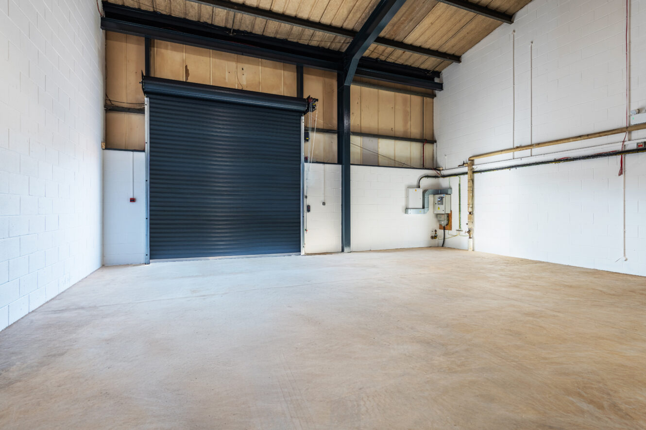 Empty industrial warehouse interior with white brick walls, exposed ceiling beams, concrete floor, a black roller shutter door, and visible electrical panels on the right wall.