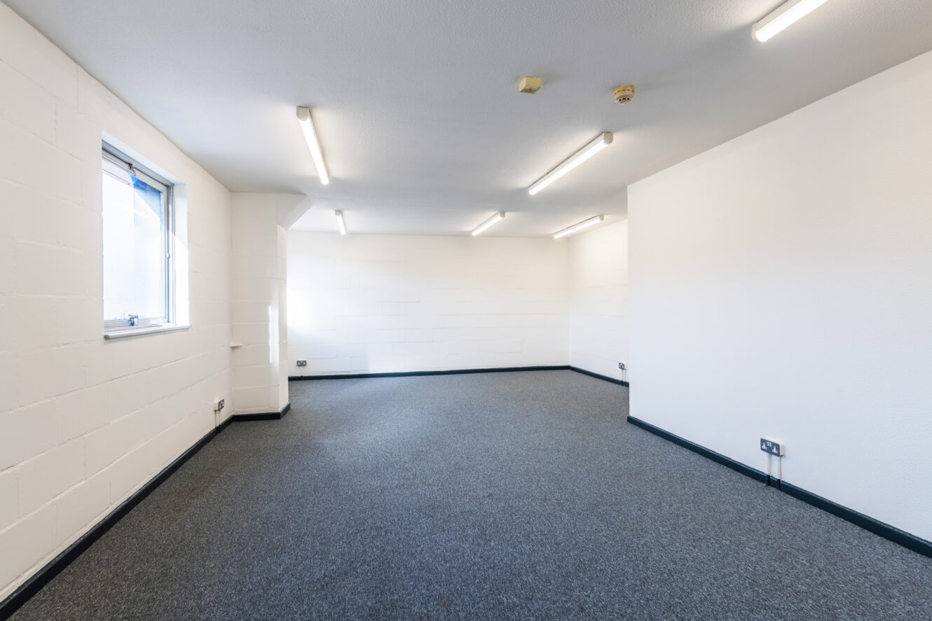Empty office space with white walls, grey carpet, ceiling lights, electrical outlets, and a single window on the left wall.