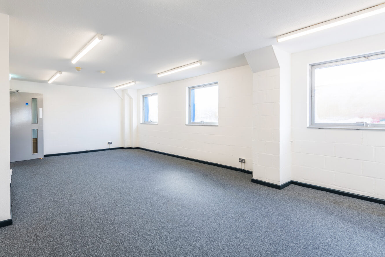 Unfurnished office space with white walls, grey carpet, three windows, ceiling lights, and a closed door.