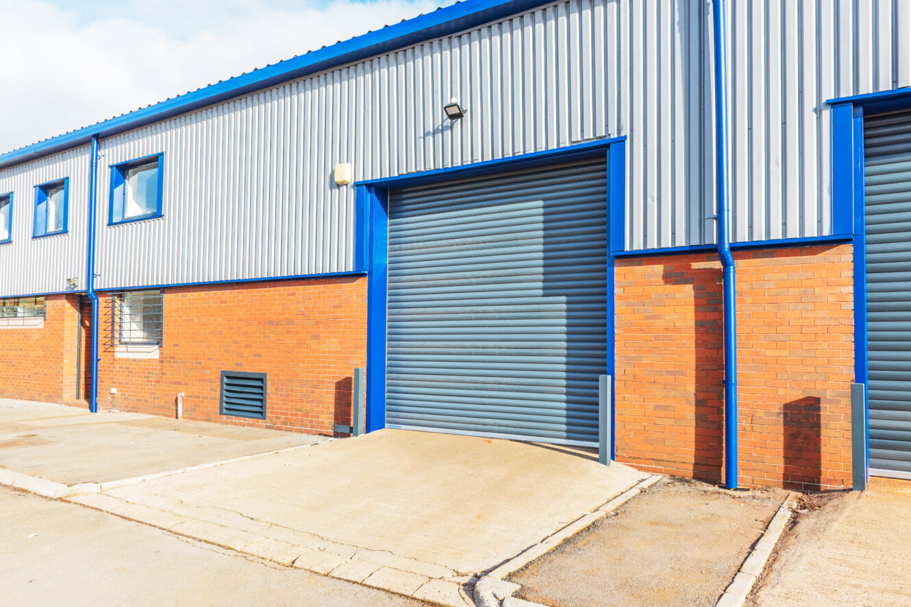 A modern industrial warehouse exterior with a blue and gray roller shutter door, brick lower facade, and metal upper panels.