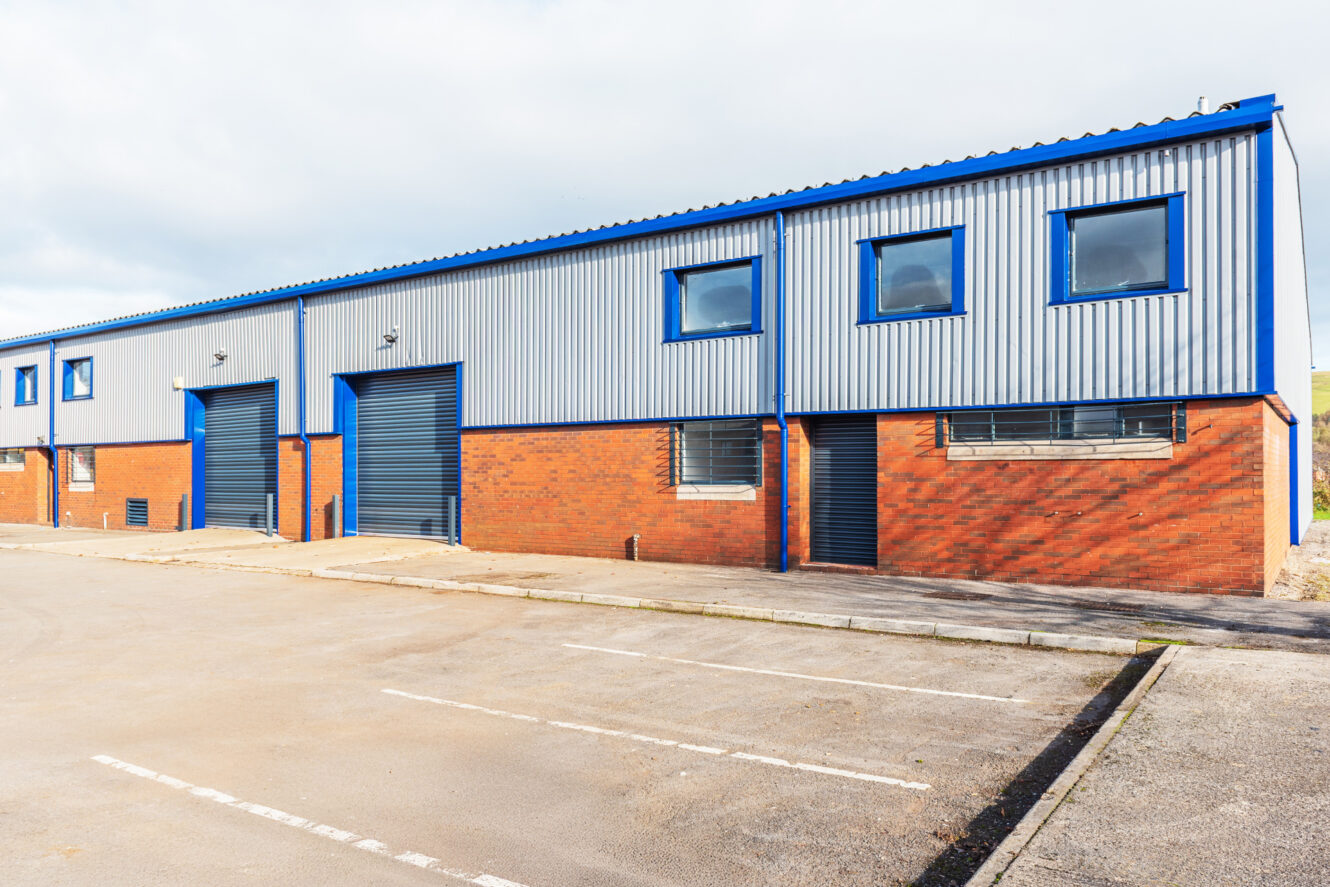 Exterior view of a modern industrial warehouse building with blue doors, metal siding, and an empty parking lot in front.