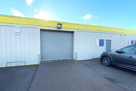 A grey metal industrial building with a closed roller shutter door, yellow trim along the roof, and a parked car on the right.