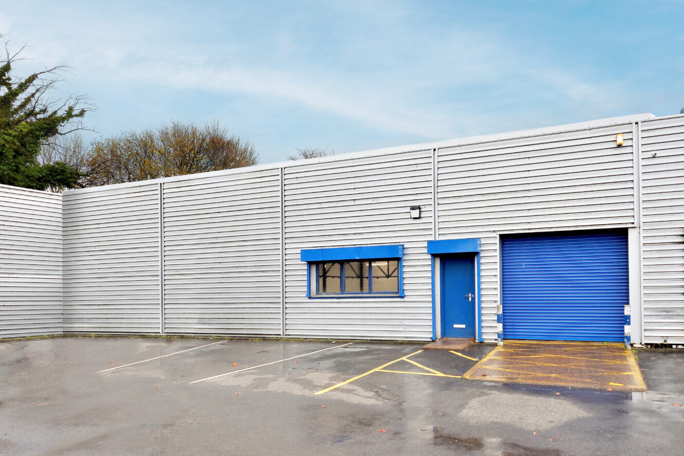 A gray industrial warehouse exterior with a blue door, blue window awning, blue roller shutter, and empty parking spaces on a wet surface.