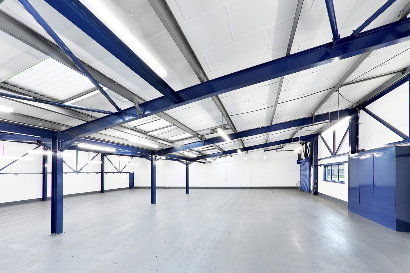 Spacious, empty industrial warehouse interior with blue steel beams, white walls, large windows, and a gray concrete floor.