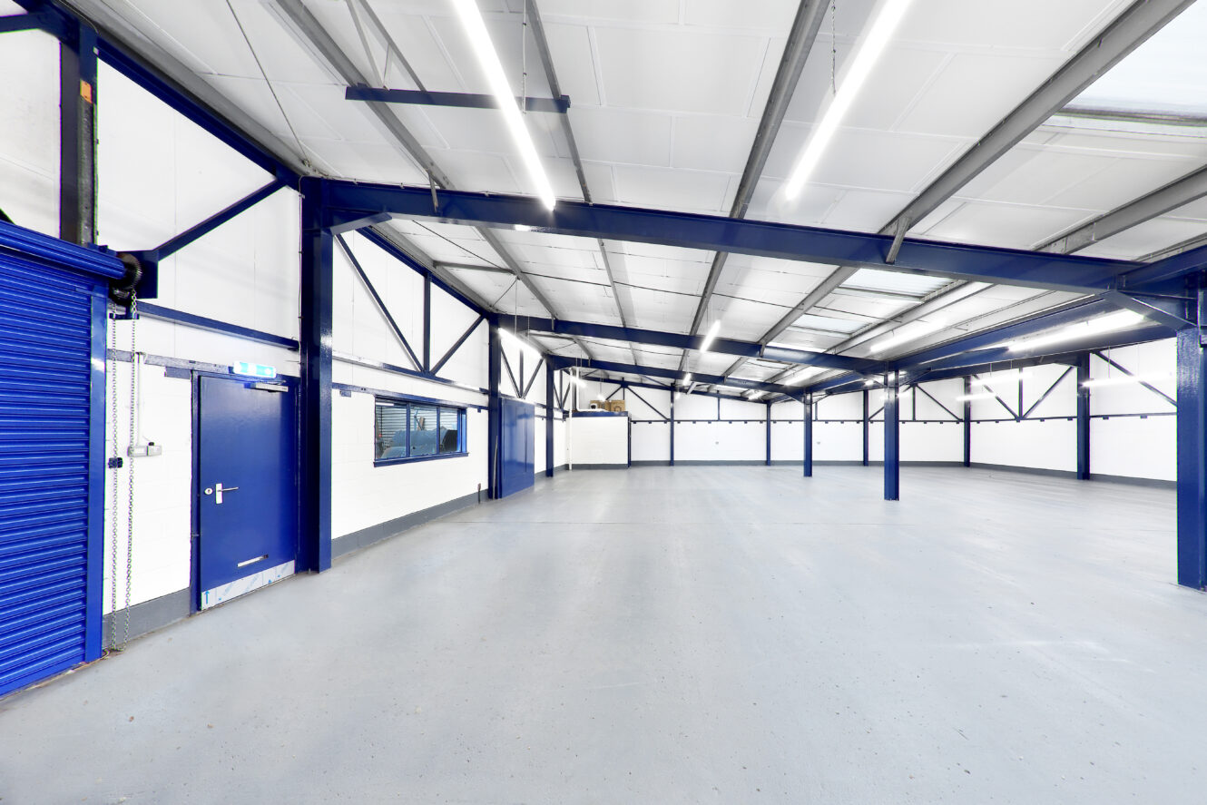 Spacious empty warehouse with blue steel beams, white walls, grey floor, fluorescent ceiling lights, and a closed blue shutter door on the left.