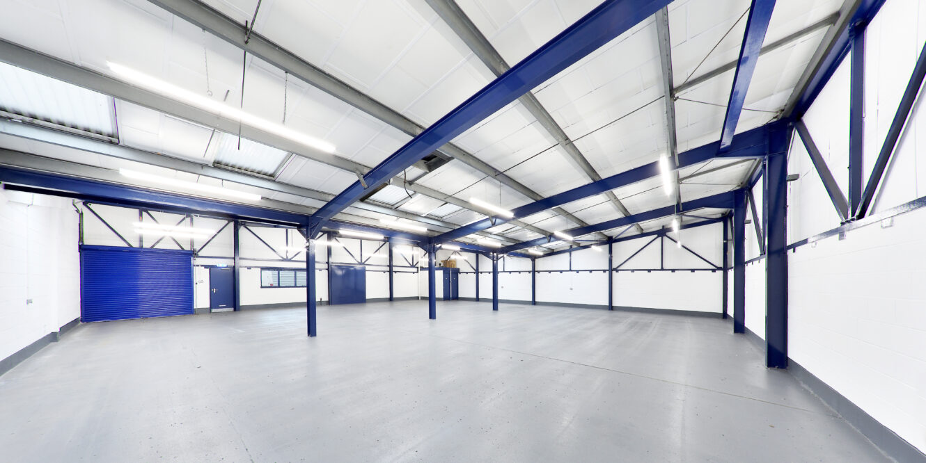 Large empty warehouse interior with white walls, blue steel beams, a grey concrete floor, and fluorescent ceiling lights. A blue roller door and windows are visible on the left wall.