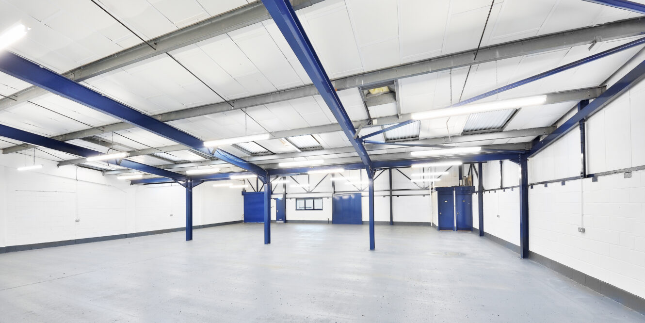 Empty industrial warehouse with white walls, exposed blue steel beams, gray concrete floor, and fluorescent ceiling lights.