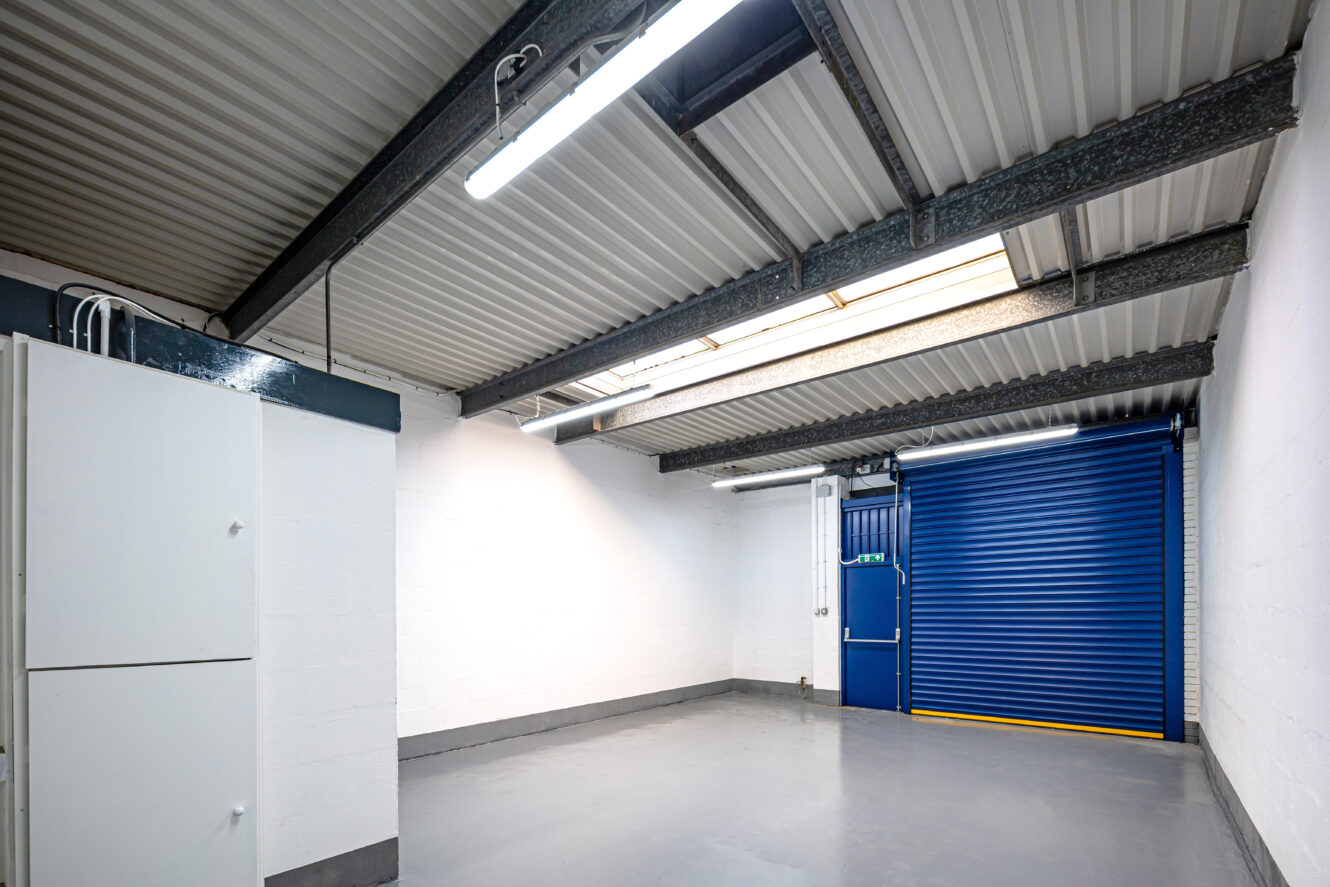 Clean, empty garage with white walls, grey floor, exposed beams, ceiling lights, and a blue roller shutter door partially open to the outside. White storage cabinet on the left wall.