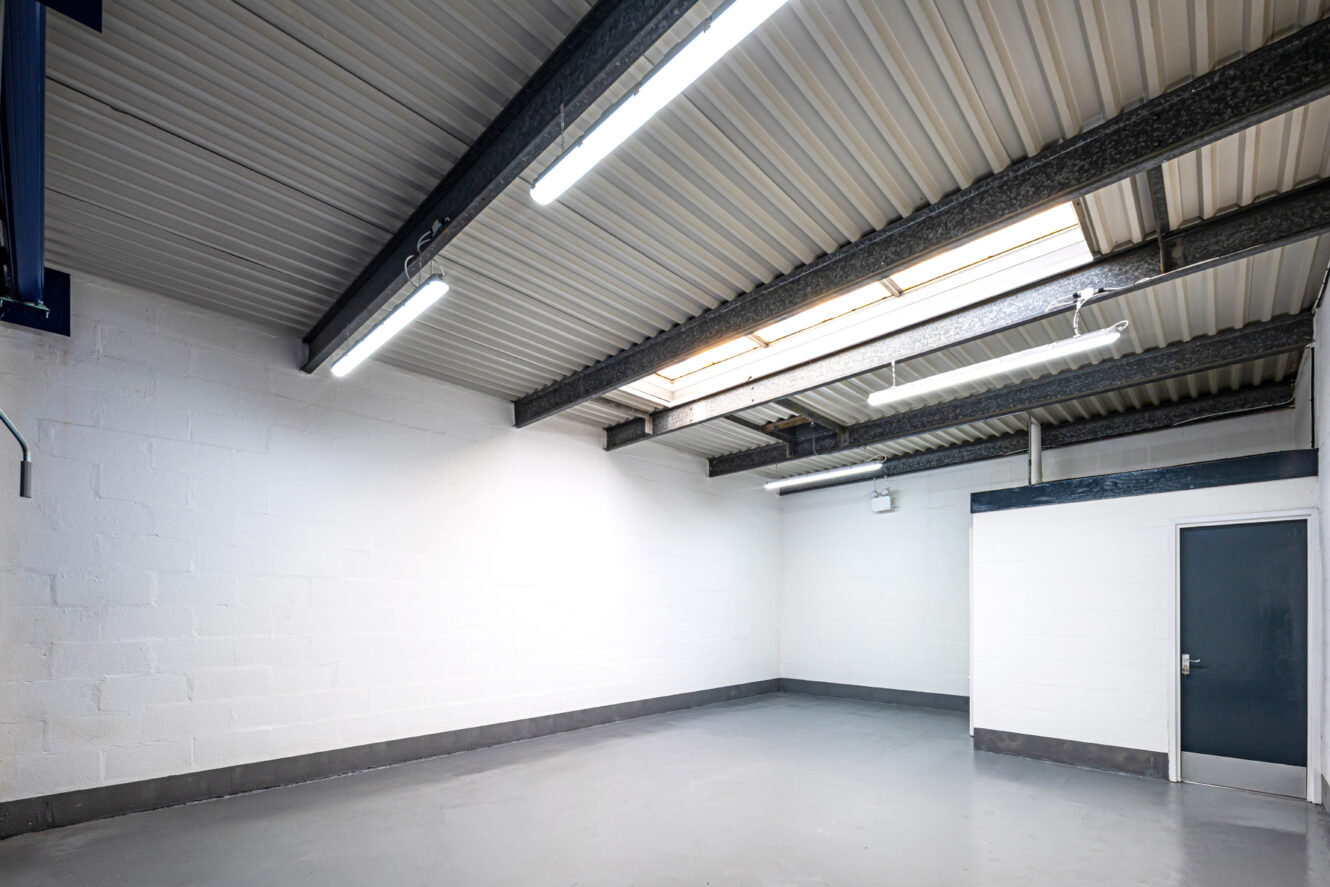 Empty industrial-style room with white painted brick walls, concrete floor, exposed ceiling beams, fluorescent lights, a skylight, and a closed gray door.