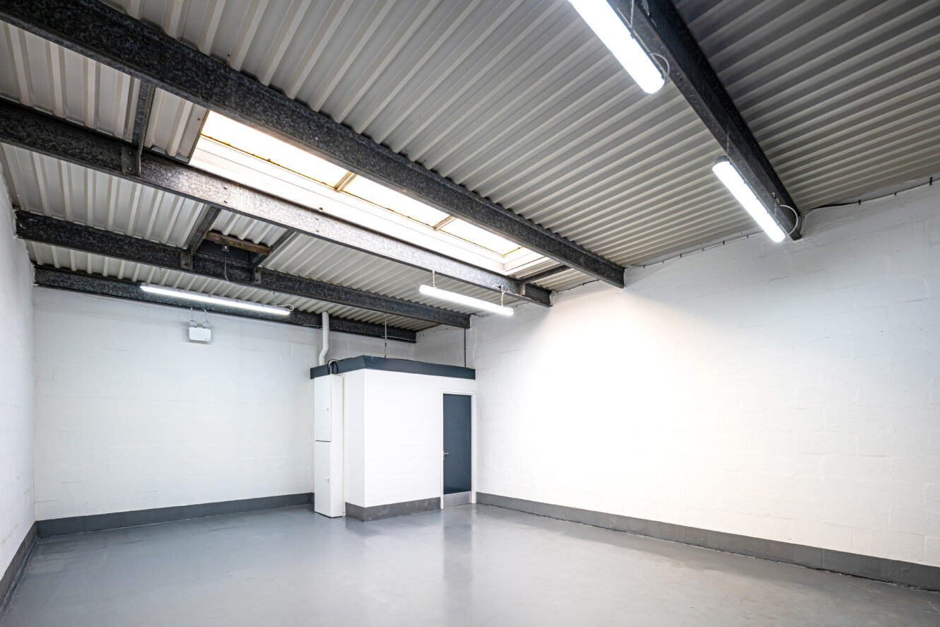 An empty industrial room with white walls, a gray concrete floor, exposed beam ceiling, fluorescent lights, and a small enclosed area with a door.