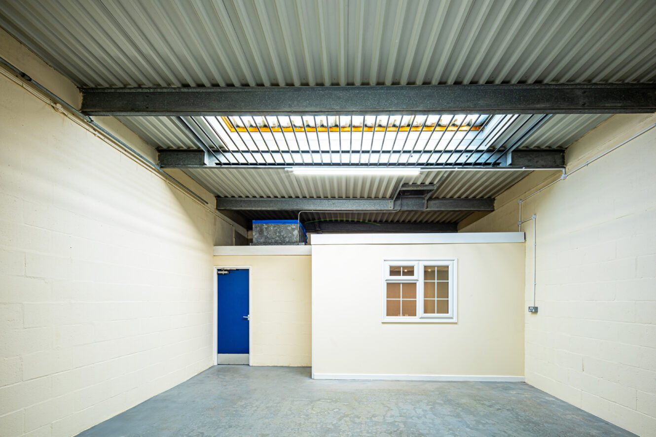 Empty industrial room with high corrugated ceiling, cream walls, concrete floor, and a small interior office with a window and a blue door.
