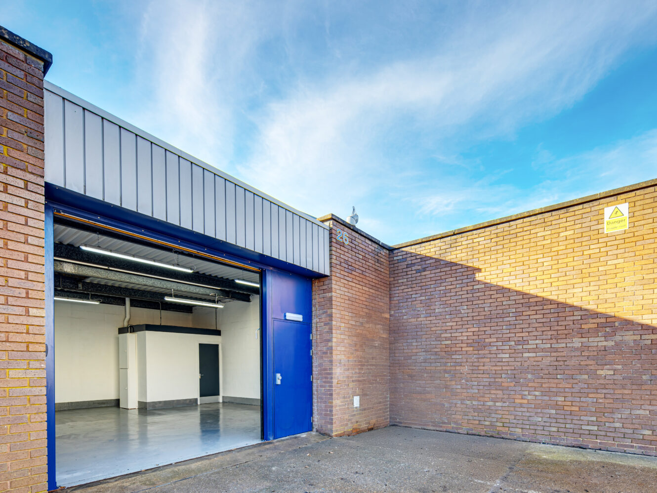 A small industrial unit with an open blue roller shutter door, exposing a clean, empty interior; brick walls and a clear sky are visible outside.