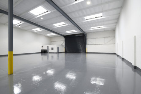 Empty industrial warehouse with glossy gray floor, high ceilings, white walls, a large black roller door, skylights, and a small enclosed office space in the corner.
