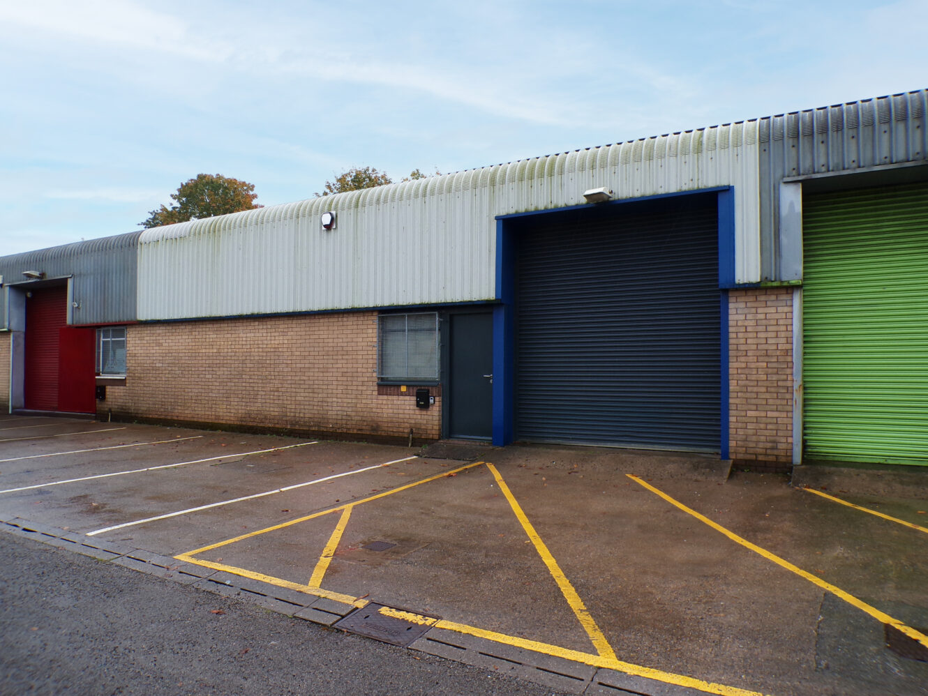 An industrial building with three colored roller shutter doors—red, blue, and green—next to empty parking spaces marked with yellow lines.