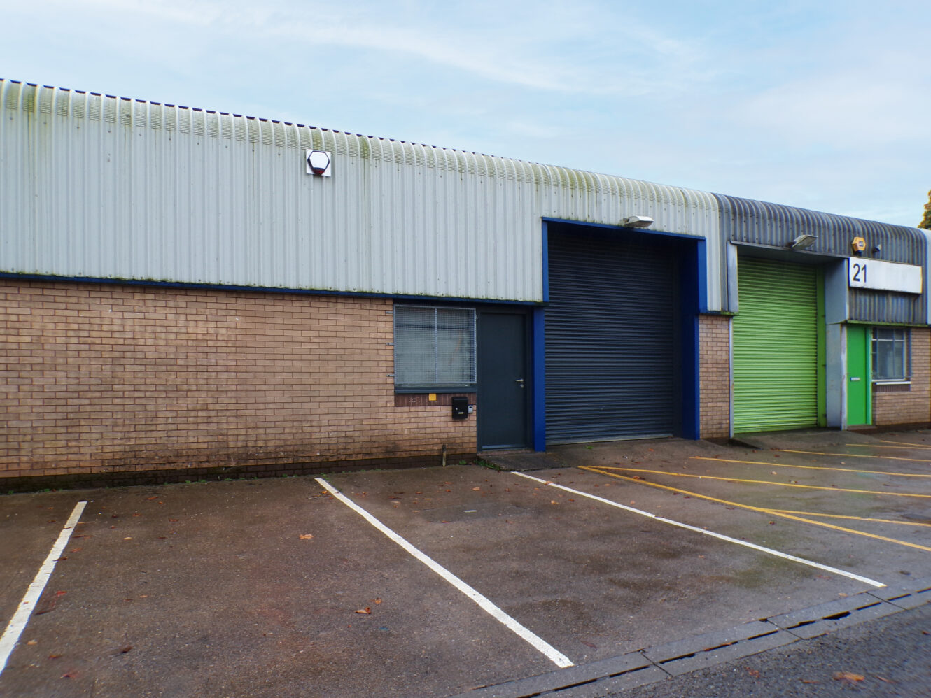 A row of industrial units with closed shutters, one blue and one green, numbered 21, with empty parking spaces in front on a wet pavement.