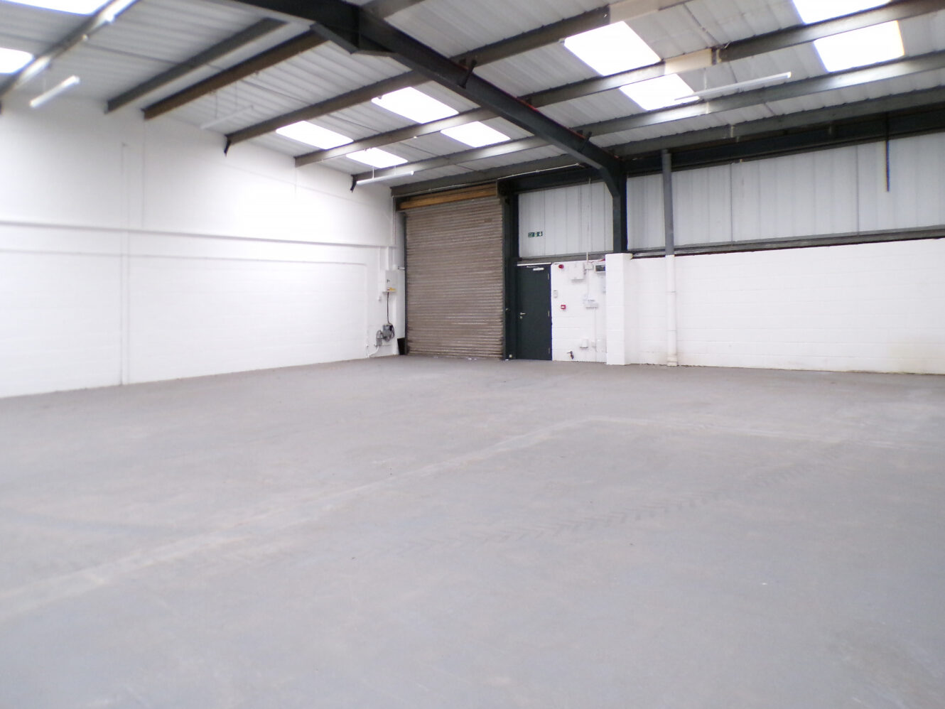 Empty industrial warehouse with concrete floor, white walls, high ceiling, skylights, and a large metal roller door at the back.