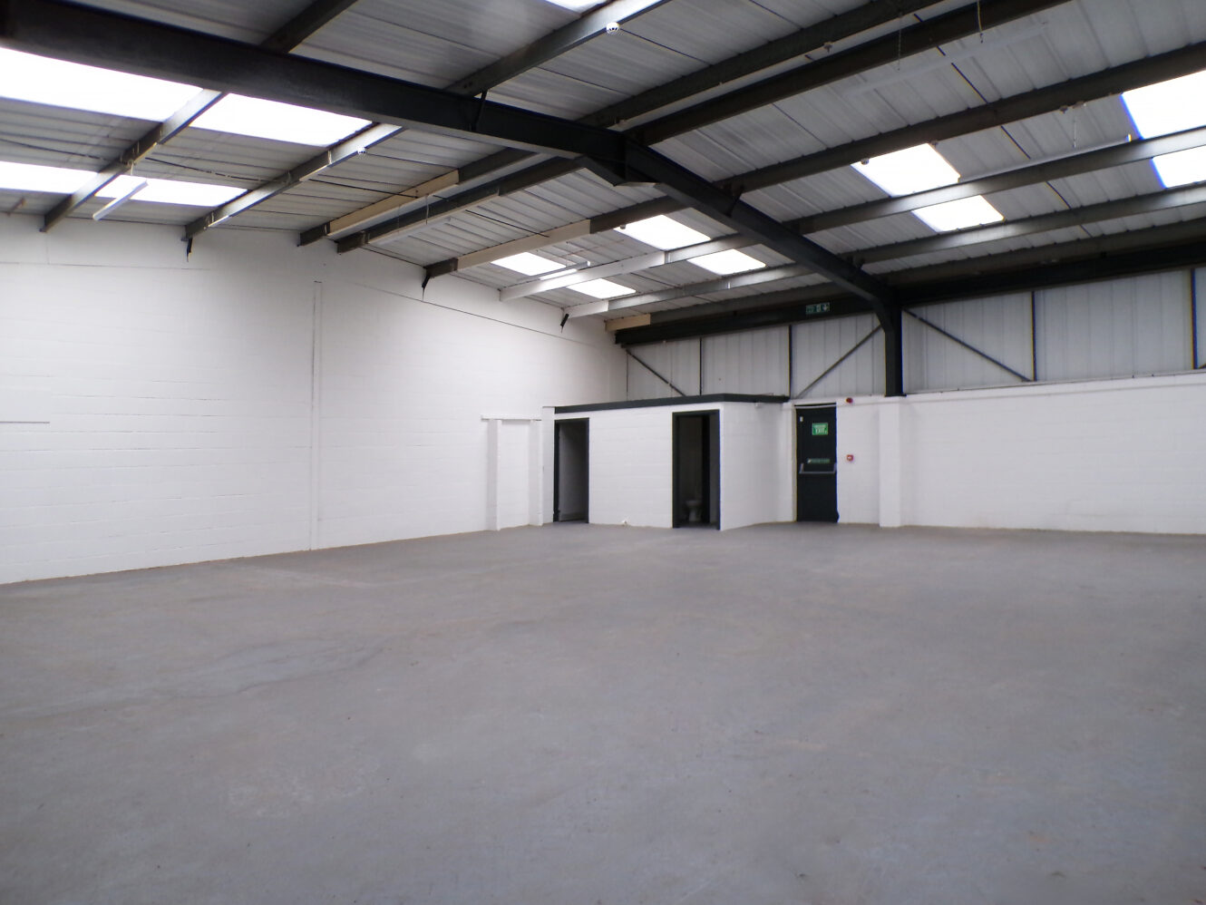 Empty industrial warehouse with concrete floor, white painted brick walls, and metal roof; two doorways and one fire exit visible on the far wall.