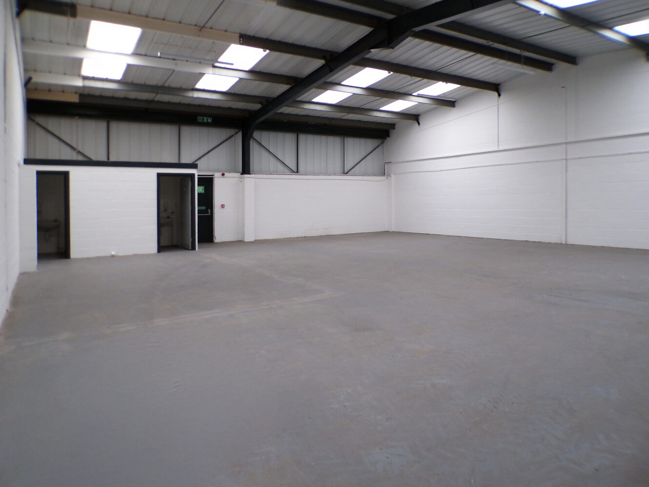 Interior of an empty warehouse with white walls, grey flooring, exposed beams, and skylights in the ceiling; two open doorways are visible on the left wall.