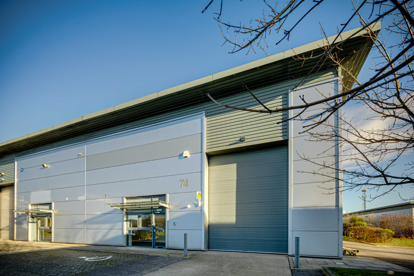 Modern industrial warehouse building with large roller door, labeled 74, against a clear blue sky and surrounded by paved area and sparse landscaping.