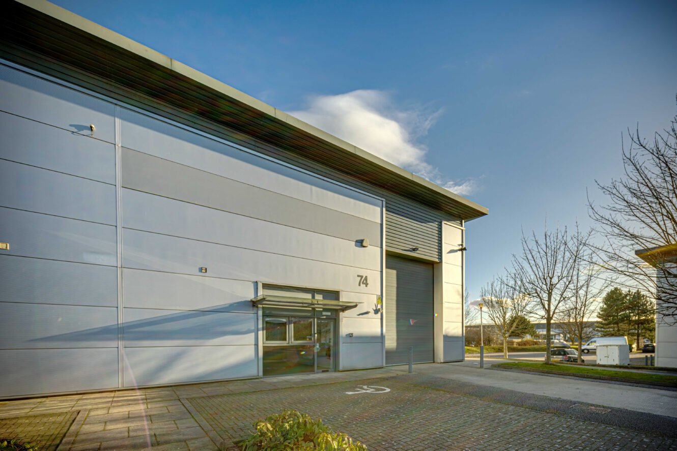 Modern industrial building with a large gray exterior, glass entrance, and numbered 74 on the wall, surrounded by a paved area and some trees under a clear sky.
