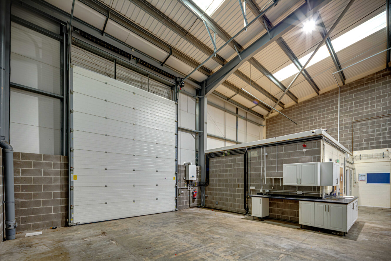 Interior of an empty industrial warehouse showing a large roller shutter door, concrete floor, brick walls, and a small office area with cabinets and electrical panels.