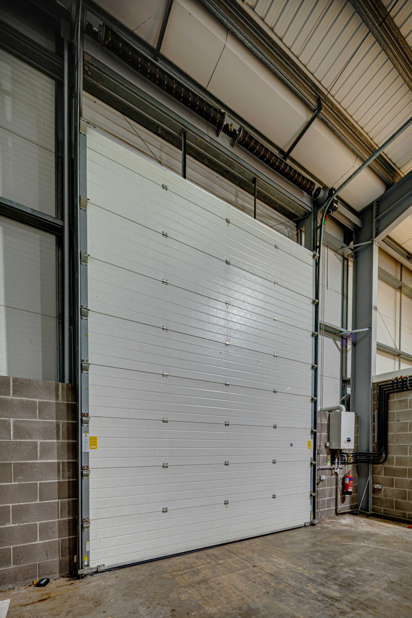 Large white industrial roll-up garage door closed inside a warehouse with concrete block walls and exposed steel beams.