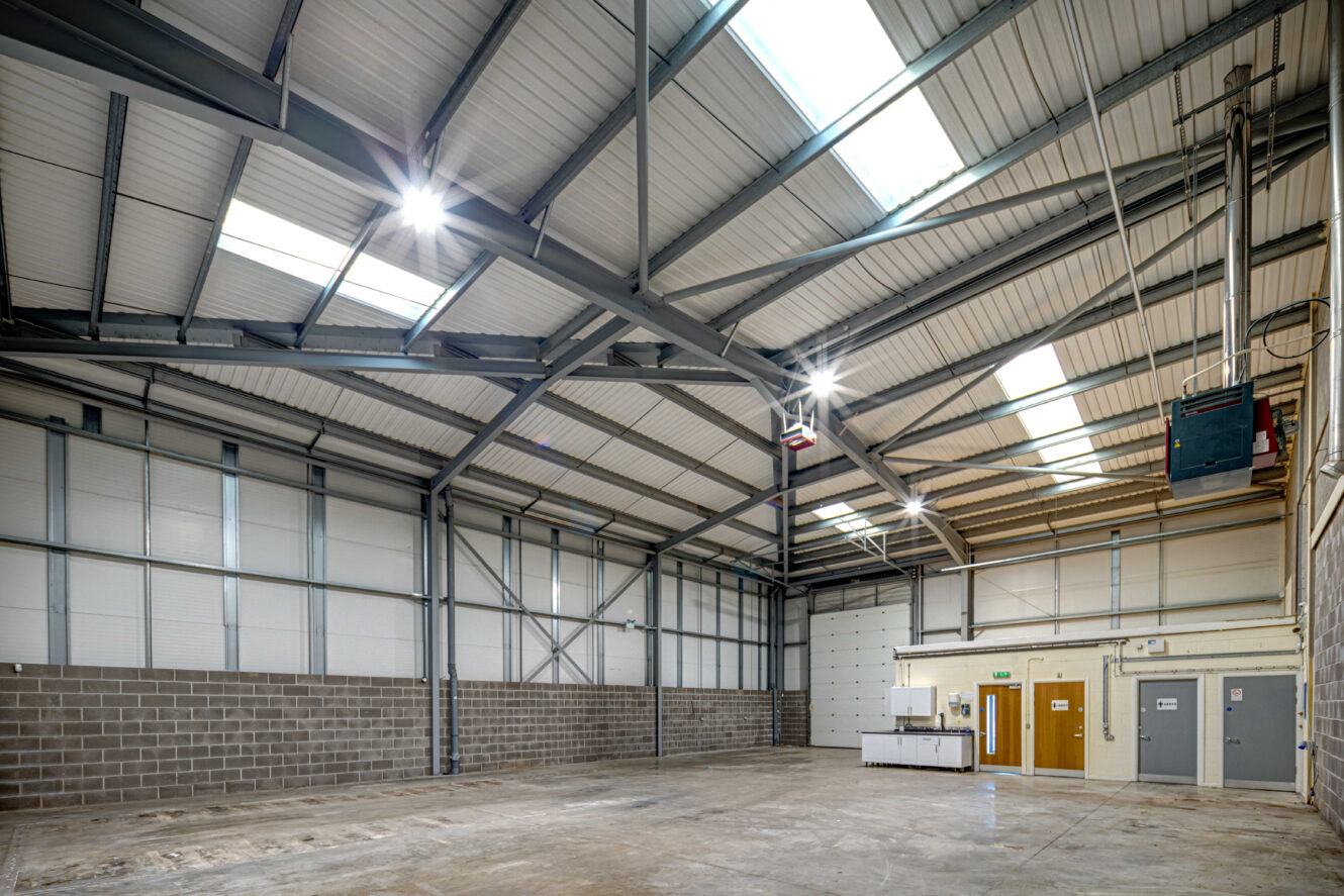 Interior of an empty industrial warehouse with high metal ceiling, concrete floor, brick lower walls, and a small office area with doors and a kitchenette. Brightly lit by ceiling lights.