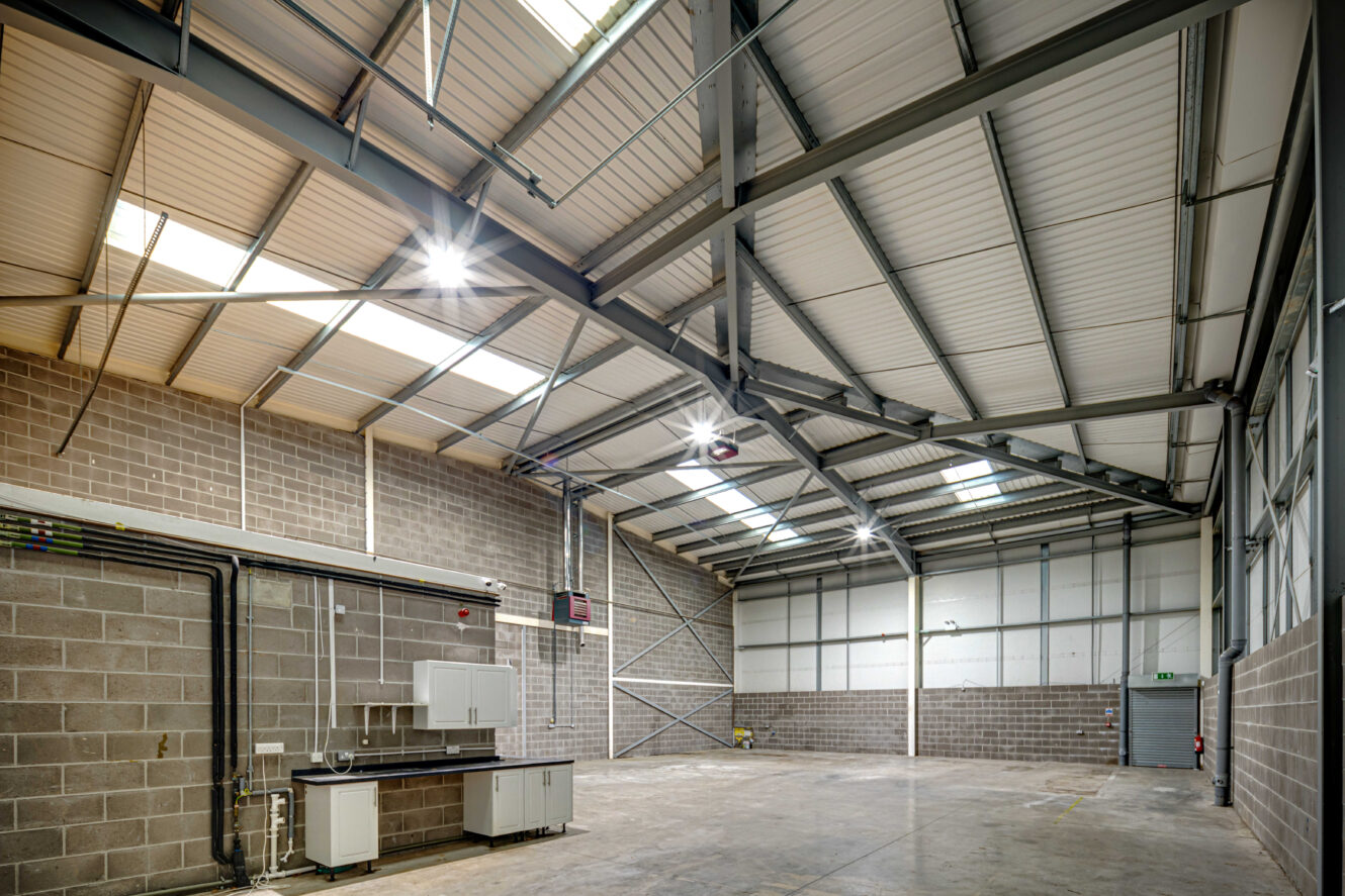 Interior of an empty industrial warehouse with exposed steel beams, brick walls, concrete floor, skylights, and a small utility area with cabinets and a sink.