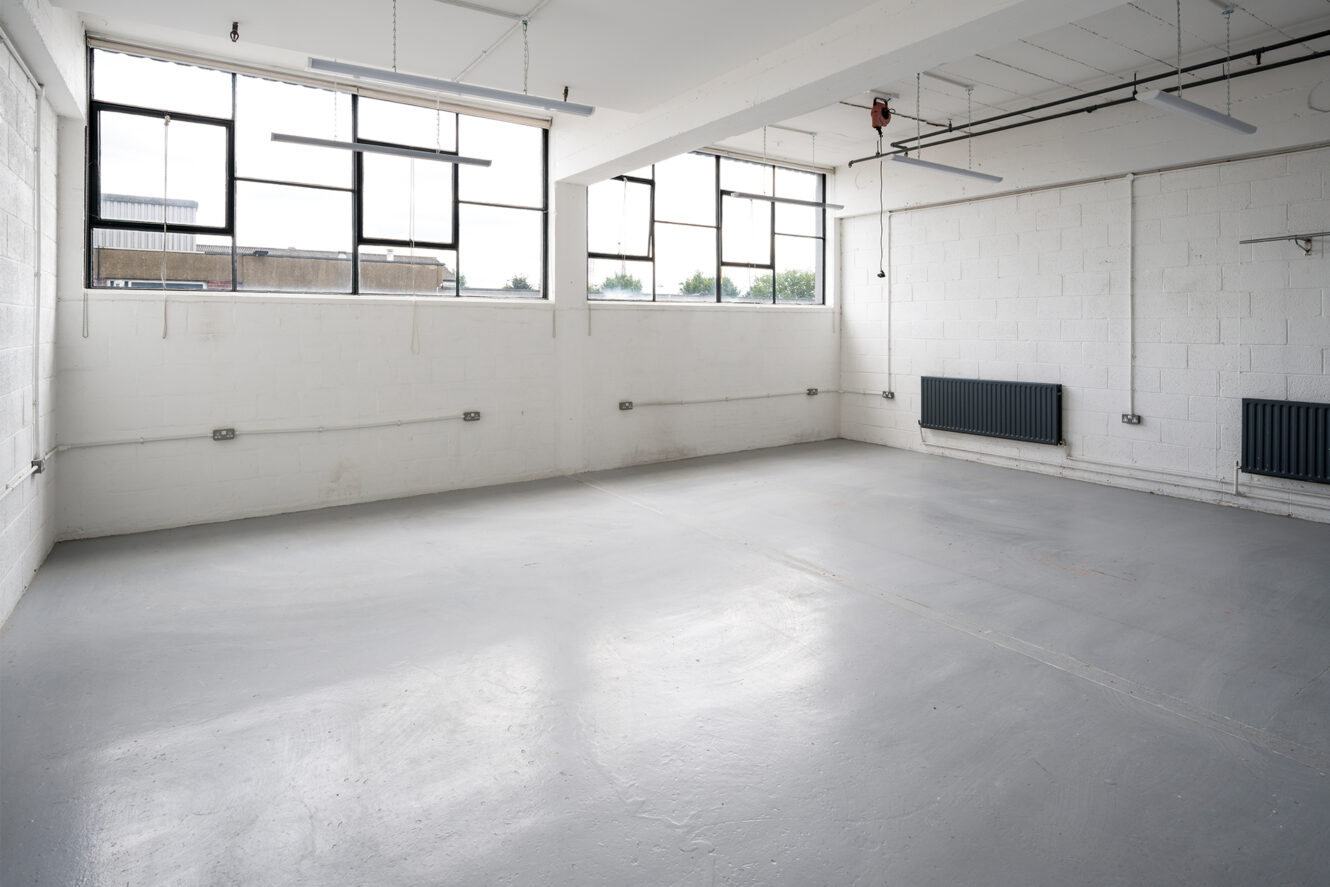 Empty industrial room with white brick walls, large windows, exposed ceiling fixtures, wall-mounted radiators, and a bare concrete floor.