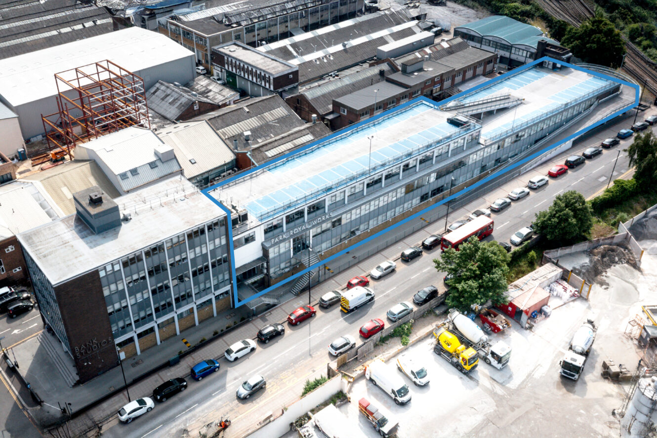 Aerial view of a large multi-story industrial building with a blue roof, adjacent to a street with parked cars and trucks, and surrounded by other warehouses and construction sites.