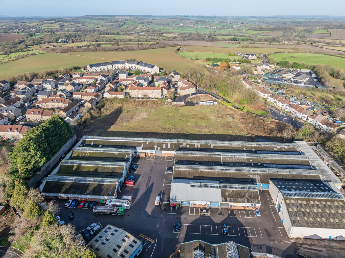 Aerial view of industrial buildings with parked vehicles in the foreground, residential houses and fields in the background under a clear sky.