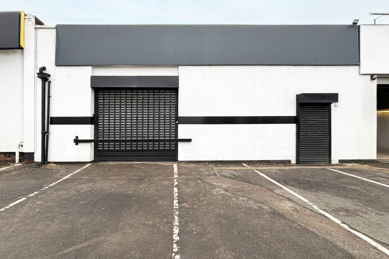 A blank commercial building facade with a closed roller shutter door, a smaller side door, and empty parking spaces in front.