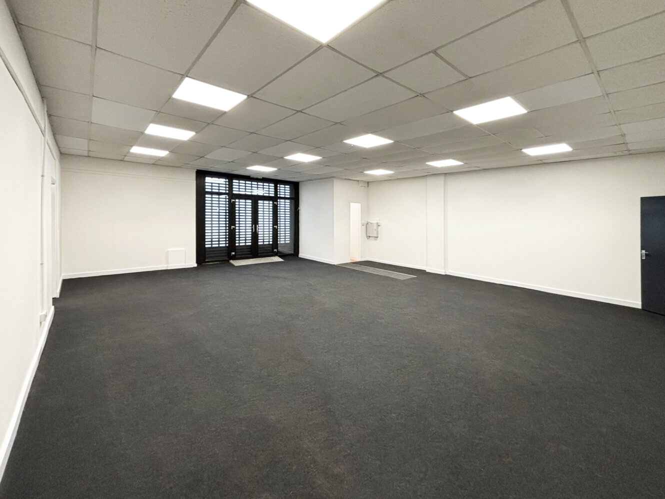 Large empty room with white walls, black carpet flooring, ceiling lights, a dark door, and large windows with blinds at the far end.
