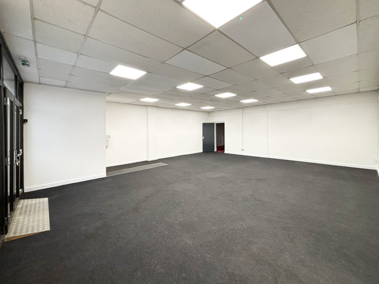 Empty office space with white walls, a gray carpet, ceiling lights, and a single closed door on the far wall.