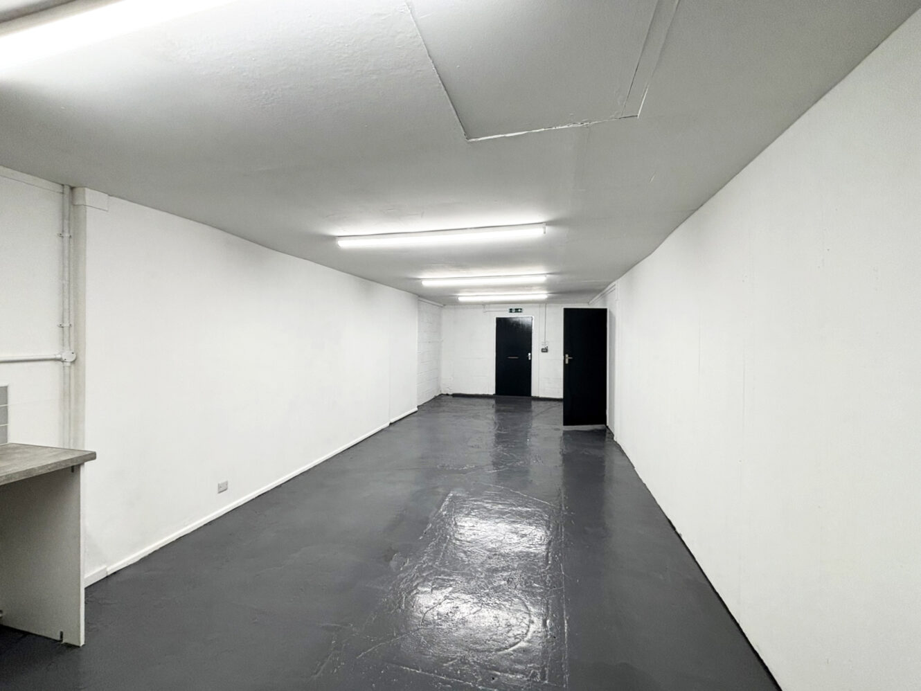 A long, empty room with white walls, a gray floor, fluorescent ceiling lights, and two black doors at the far end.